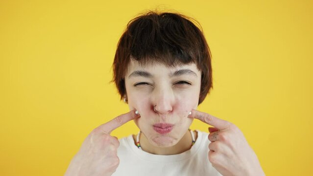 Cute short-haired skinny young adult caucasian girl looking at camera, doing duck face, and pointing at her dimples over yellow background. High quality 4k footage