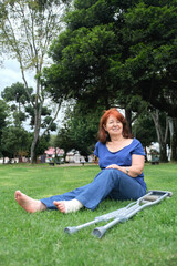 Mature woman resting on the grass, smiling, barefoot, with a bandage on her left foot and crutches at her side. Concepts: positive attitude towards an injury, recovery and physical rehabilitation.