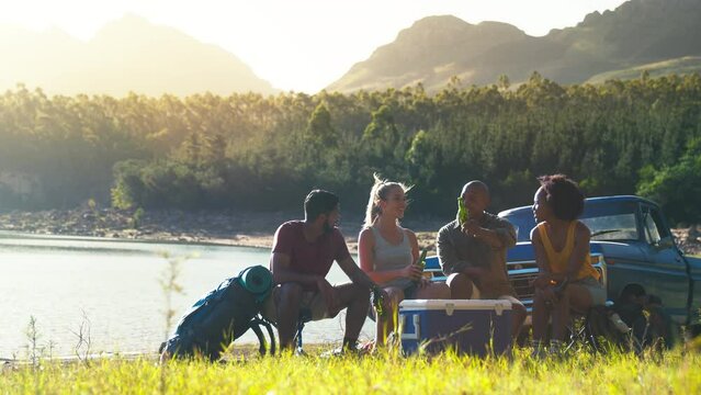 Group of friends with backpacks by pick up truck on road trip drinking beer with cheers from cooler by lake - shot in slow motion