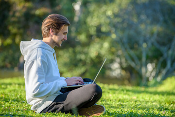 Obraz premium Happy Student with laptop sits on green lawn. Young smiling guy is chatting online with friends in the park. Cheerful Young man freelancer works remotely at a computer on the Internet outdoors.
