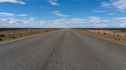 Fototapeta na wymiar A long straight road without lines that crosses the Atacama desert in Chile. Sun with clouds in the blue sky.