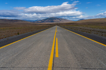 A long straight road without lines that crosses the Atacama desert in Chile. Sun with clouds in the blue sky.