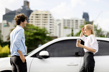 Progressive businessman and businesswoman with coffee, standing at electric car connected to...