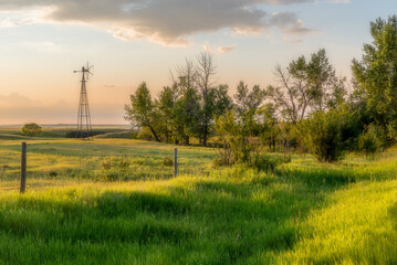 A pastoral scene of an old windmill in a pasture at sunset with trees and a fence line in the foreground- Orton Effect