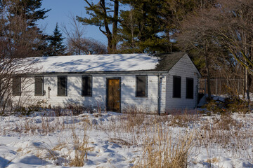 Abandoned house - windows blocked, electricity wires cut, snow covered field with trees in the background. Spring, sunny day.