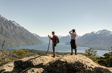 amigos sobre una roca en la cima de un cerro sacando fotos y mirando el paisaje de montañas nevadas y lagos
