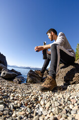 chico mirando el paisaje sentado a la orilla rocosa del lago tomando bebiendo mate argentino con...