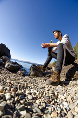 chico mirando el paisaje sentado a la orilla rocosa del lago tomando bebiendo mate argentino con montañas de fondo