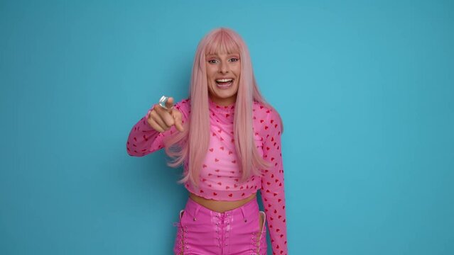 Pretty girl in pink top and with pink hair stands isolated in the middle of blue studio background, points finger directly to the camera urging to take advantage of discounts, sale discounts concept