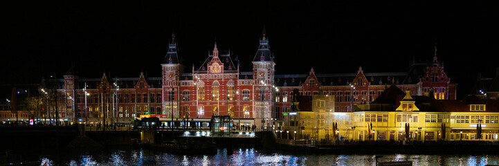 Obraz premium Amsterdam Centraal buildings, water reflections, and busy street at night in Netherlands, vibrant long exposure photography