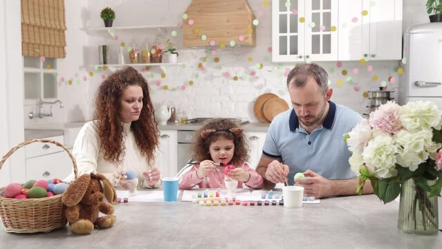Portrait of cute baby girl helping her parents to paint Easter eggs, while wearing bunny ears. Charming family having good time together. High quality 4k footage