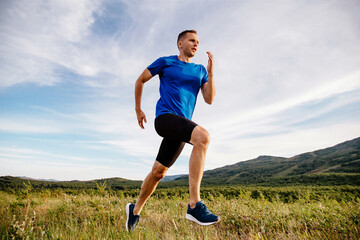 sporty male runner running on mountain trail in summer, front view, blue shirt and black half tights, sports photo