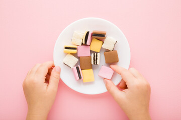 Baby girl hand taking colorful jelly gummy candies from white plate on light pink table background. Pastel color. Children sweets. Closeup. Point of view shot. Top down view.