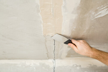 Young adult man hand using spatula and plastering concrete ceiling with putty on mesh in electric wire channel. Closeup. Repair work of home. Renovation process.