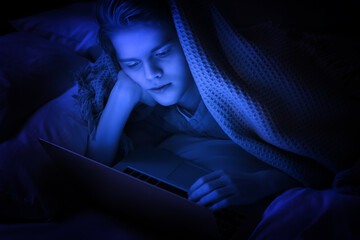 Internet addiction. Teenage boy using laptop under blanket on bed at night. Toned in blue © New Africa