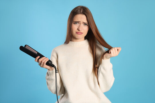 Upset young woman with flattening iron on light blue background. Hair damage