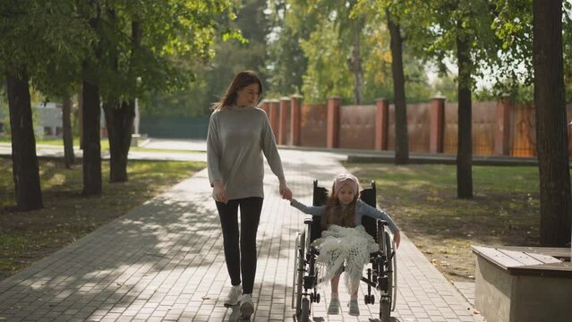 Smiling mother and daughter with paralysis in wheelchair enjoy walk in park. Woman spends time with little girl on fresh air on sunny spring