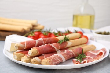 Plate of delicious grissini sticks with prosciutto, cheese and rosemary on light grey table, closeup