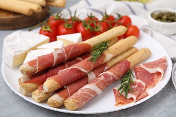 Plate of delicious grissini sticks with prosciutto, cheese and tomatoes on light grey table, closeup