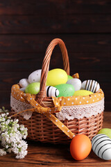 Wicker basket with festively decorated Easter eggs and white lilac flowers on wooden table