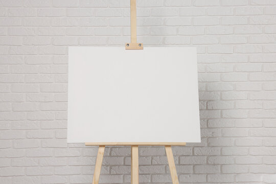Wooden easel with blank canvas near white brick wall. Space for text