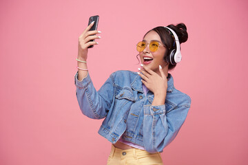 Young Asian teen woman using smart phone she listening music in headphones isolated on pink background.