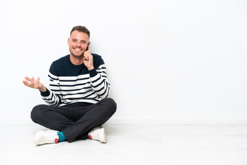 Young man sitting on the floor isolated on white background keeping a conversation with the mobile phone with someone
