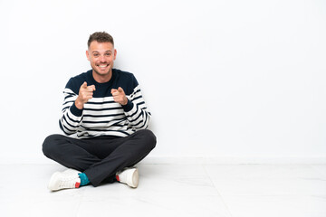Young man sitting on the floor isolated on white background pointing to the front and smiling