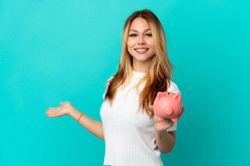 Teenager blonde girl holding a piggybank over isolated blue background extending hands to the side...