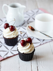 two cupcakes with white cream and cherry on a light wooden table