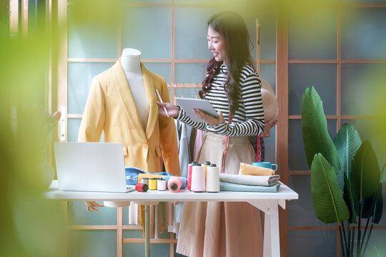 Young women fashion designer using laptop working and received order customer for design tailor and cloth in the showroom,  Lifestyle Stylish tailor taking measurements on mannequin for cutting cloths