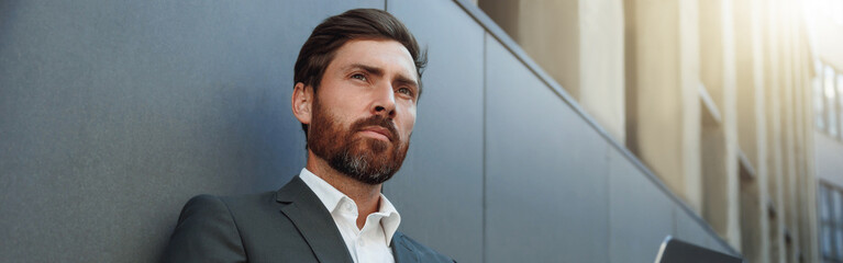 Businessman working on laptop outside office building and looking at side