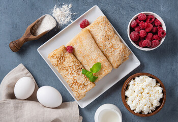 Homemade crepes with raspberry and cottage cheese on a plate on a blue background with eggs and flour. Healthy food concept.