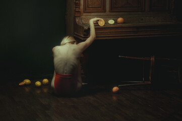 A girl sits on the floor and picks up an orange from the table. View from the back. A red dress, a piano, an overturned chair, and fruit. Dark interior. Artistic style