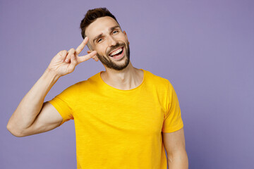 Young smiling cheerful joyful cool fun happy caucasian man wear yellow t-shirt showing cover eye with victory sign isolated on plain pastel light purple background studio portrait. Lifestyle concept.