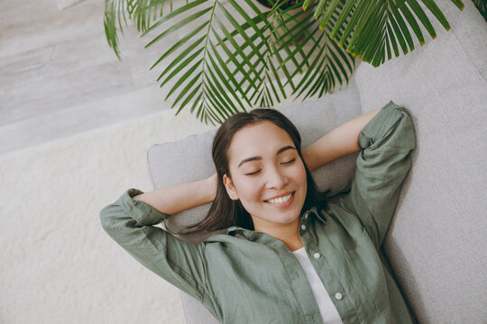 Top view young woman of Asian ethnicity wears casual clothes hold hands behind neck close eyes sits on grey sofa couch stay at home hotel flat rest relax spend free spare time in living room indoor.
