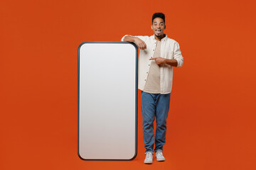 Full body young man wearing light shirt casual clothes point index finger on big huge blank screen mobile cell phone smartphone with workspace copy space mockup area isolated on orange red background.