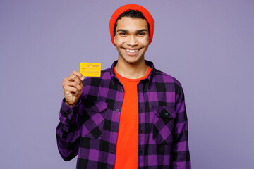 Young smiling happy fun man of African American ethnicity wear casual shirt orange hat hold in hand mock up of credit bank card isolated on plain pastel light purple color background studio portrait.