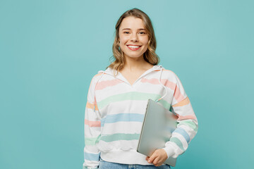 Smiling student cheerful happy young IT woman wears striped hoody hold closed laptop pc computer looking aside isolated on plain pastel light blue cyan background studio portrait. Lifestyle concept.