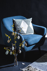 interior, holidays and home decor concept - close up of modern blue chair with pillow and easter eggs hanging on forsythia branches in vase on floor in dark room