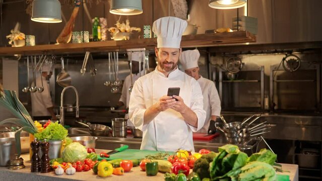 Portrait of attractive bearded caucasian male cook using smartphone online searching for recipe before cooking. Beautiful skillful famous chef using technologies during cooking food.
