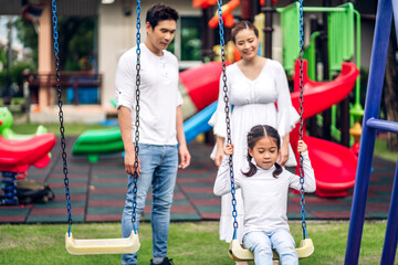 Portrait of enjoy happy love asian family father and mother with little asian girl smiling playing and pushing daughter on the swing moments good time at playground