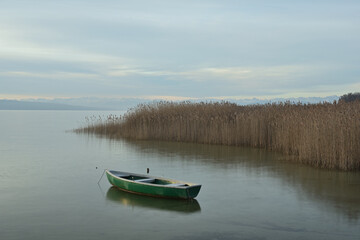 Eine morgendliche Stimmung an einem See, in dem ein kleines Boot treibt, und hinter einer Schilffläche sie das man das Bergpanorama