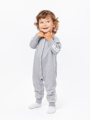 A 2-year-old toddler with curly hair in a gray jumpsuit and socks makes a wish, dreams, folding his hands on a white background.