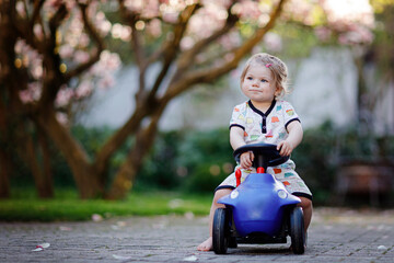 Cute little baby girl playing with blue small toy car in garden of home or nursery. Adorable...