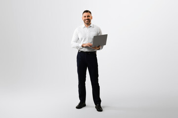 Full length portrait of middle aged businessman using laptop computer for online work, grey studio...