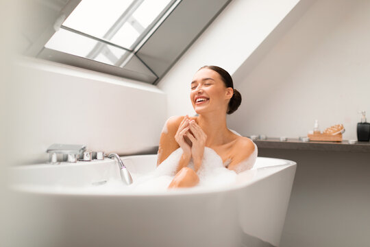Woman Sitting In Bathtub Taking Bath With Foam In Bathroom