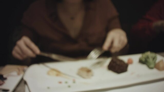 still view of blurry view of a woman wearing dark brown dress holding fork and knife eating food of steak vegetable on a huge long rectangle white plate in a restaurant