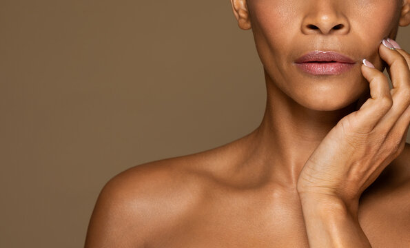 Cropped of black middle aged woman touching cheek, demonstrating perfect skin, brown background, free space