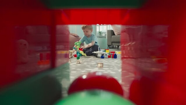Girl holds baby doll while boy playing with robot dinosaur. Kids sit on floor near handmade wooden house view through clearance in construction of cubes